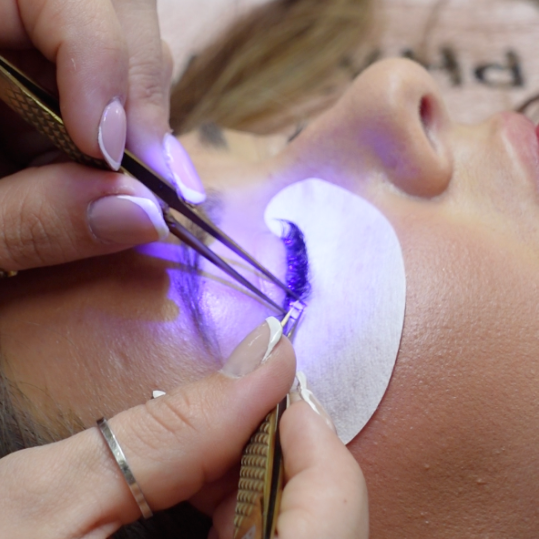 Close-up of a lash extension application in progress, with a client lying down and a technician using tweezers to place a single black lash with UV Lash Glue.